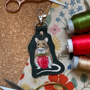 May include: A green leather keychain with an embroidered image of a brown mouse holding a red strawberry. The keychain has a silver metal ring.