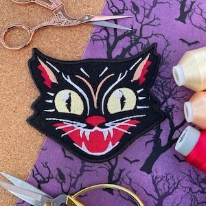 May include: Black felt iron-on patch of a cartoon cat with large yellow eyes, a red mouth with sharp teeth, and white whiskers. The cat's fur is black with a white stripe down the nose.