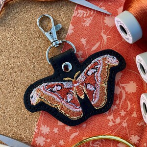 May include: A black leather keychain with a red and white embroidered moth design. The keychain has a silver metal clasp.