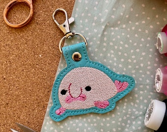 May include: A teal blue key chain with a pink and white embroidered axolotl design. The axolotl has pink gills and black eyes.
