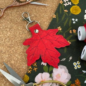 May include: A red maple leaf keyring with a silver clasp. The leaf is made of textured material and has detailed vein patterns. The background includes scissors, floral fabric, and spools of thread.