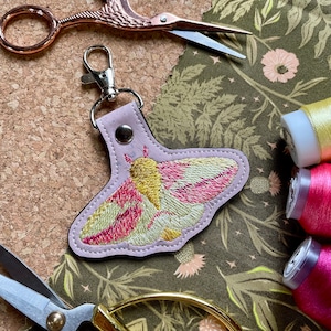 May include: A pink leather keychain featuring an embroidered yellow and pink moth design. The keychain is displayed on a green floral fabric background.