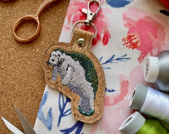 May include: A cork keychain featuring an embroidered manatee with green and gray details. The keychain is attached to a silver key ring.