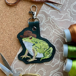 May include: A green frog wearing a brown cowboy hat is embroidered on a dark green leather keyring. The keyring has a silver metal clasp.