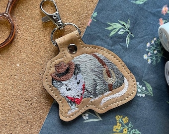 May include: A brown cork keychain with a white and gray embroidered possum wearing a brown cowboy hat and a red bandana. The possum is holding a lasso in its right paw.