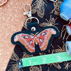 May include: A black leather keychain with a red and white embroidered butterfly design. The keychain is attached to a silver key ring. The keychain is about 2 inches long.