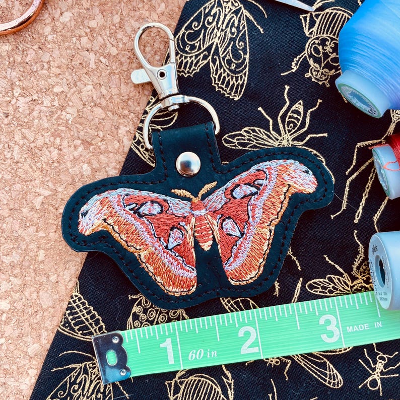 May include: A black leather keychain with a red and white embroidered butterfly design. The keychain is attached to a silver key ring. The keychain is about 2 inches long.