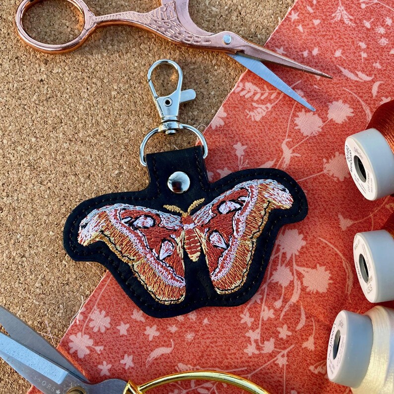 May include: A black leather keychain with an embroidered moth design. The moth has orange, white, and brown wings. The keychain has a silver metal clasp.