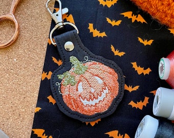 May include: A black leather key chain with a stitched embroidered pumpkin design. The pumpkin is orange with a black face and a green stem.