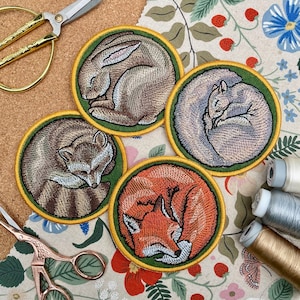 May include: Four embroidered patches featuring sleeping animals. The patches are circular and have a green outer ring with a yellow inner ring. The animals are a rabbit, a raccoon, a fox, and a squirrel. Each animal is embroidered in detail and has a realistic appearance.