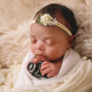 May include: A newborn baby wearing a yellow headband with a floral accent, wrapped in a cream-colored blanket, and holding a miniature black camera. The baby is sleeping peacefully on a soft, textured surface.