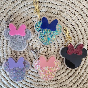 May include: Five mouse-shaped keychains with glitter and bow accents. The keychains feature a variety of colors, including silver, blue, pink, purple, and black. Each keychain has a gold-colored clasp.