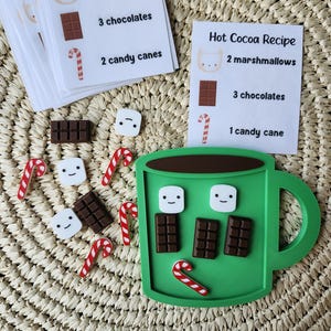 May include: A green mug-shaped board with small brown chocolate bar, white marshmallow, and red and white candy cane pieces, accompanied by printed recipe cards titled Hot Cocoa Recipe listing various quantities of these items.