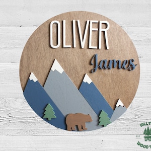 May include: A round wooden sign with a mountain scene, a brown bear, and the names "Oliver James" in white and blue lettering. The sign has a rustic wood grain and is decorated with blue, gray, and white mountains. The sign is perfect for a nursery or child's room.