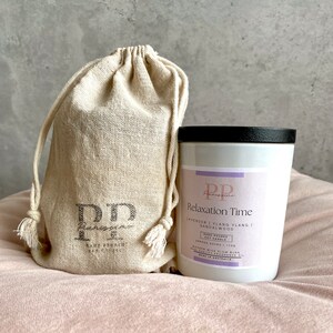 May include: A white candle with a black lid and a label that reads "Relaxation Time" with the scent notes "Lavender, Ylang Ylang, Sandalwood". The candle is hand-poured soy wax and has a wooden wick. The candle is sitting on a pink surface next to a white drawstring bag with the logo "PP" printed on it.