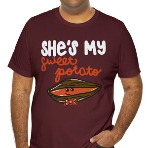 Op de afbeelding: Een maroon t-shirt met een witte afbeelding waarop staat "She's my sweet potato" met een cartoon-zoete aardappel met een strikje.