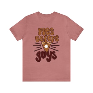 Pode incluir: Uma camiseta rosa com o texto "Pies Before Guys" em letras marrons e brancas. Uma torta marrom e branca com chantilly está no centro do texto.