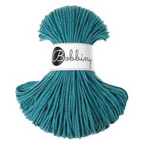 May include: A teal-colored skein of Bobbiny macrame cord. The cord is tightly wound and wrapped with a white label featuring the brand name in a stylish script. The cord is a vibrant teal color, perfect for crafting projects.