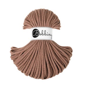 Op de afbeelding: Een bol bruin Bobbiny macramé koord. Het koord is strak opgerold en omwikkeld met een wit label met de merknaam in een sierlijk lettertype. Het koord heeft een effen, neutrale kleur, geschikt voor diverse knutselprojecten.