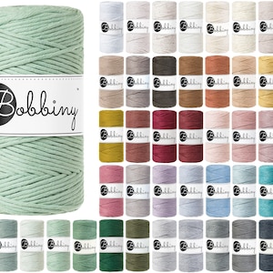 Puede incluir: Un gran carrete de cordón de macramé verde menta con una etiqueta blanca que dice "Bobbiny". Muchos carretes más pequeños de cordón de macramé en varios colores, incluyendo blanco, beige, marrón y azul, rodean el carrete grande. Para manualidades.