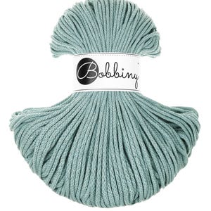 Op de afbeelding: Een streng turquoise macramé-garen. Het garen is strak opgerold en omwikkeld met een wit etiket met de tekst "Bobbiny" in een zwart, gestileerd lettertype. Het garen heeft een zachte, gedempte tint turquoise.