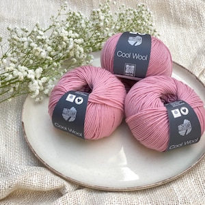 Könnte beinhalten: Drei Knäuel rosa Cool Wool-Garn liegen auf einem weißen Teller, mit weißen Blumen im Hintergrund. Jedes Knäuel hat ein schwarzes Etikett mit weißem Text. Das Garn ist ein weiches, gedämpftes Rosa.
