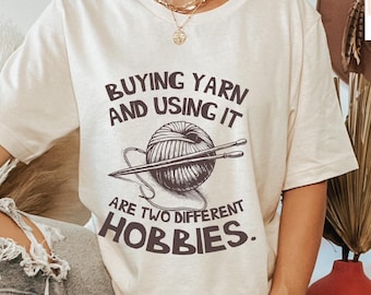 May include: A cream colored t-shirt with the text "Buying yarn and using it are two different hobbies."  The text is in brown and there is a drawing of a ball of yarn and knitting needles.