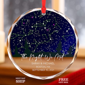 May include: A round, clear glass ornament with a red ribbon. The ornament features a dark blue night sky design with constellations and the text "The Night We Met." Below the text are names, a city, and a date.
