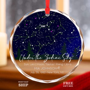 May include: A round, clear glass ornament with a red ribbon for hanging. The ornament features a dark blue background with a constellation design and the text "Under the Zodiac Sky". The ornament includes the name "MIA JOHNSON" and birth details.