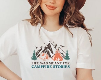 May include: White t-shirt with a graphic design of a mountain range, a campfire, a tent, and the text "Life was meant for campfire stories".