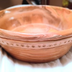 Puede incluir: Un tazón grande, redondo, de cerámica con un diseño marmoleado en tonos melocotón, marrón y blanco. El tazón tiene un borde texturizado y un interior liso.