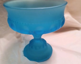 May include: A frosted blue glass pedestal bowl with a decorative design. The bowl is on a white surface.