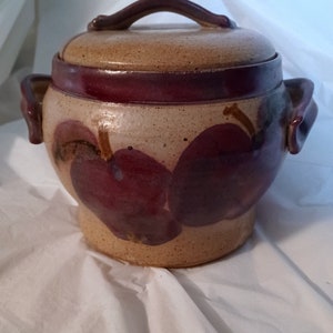 Peut inclure: Un pot en céramique avec un couvercle, orné d'un motif peint à la main de pommes violettes sur fond beige. Le pot a deux anses sur les côtés.