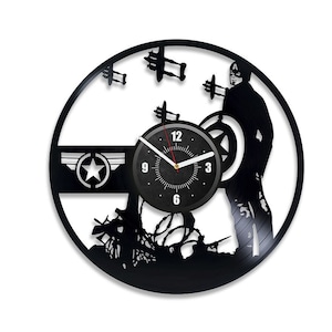 Op de afbeelding: Een zwarte vinyl platenspeler klok met een silhouet van Captain America die staat met zijn schild, een pistool vasthoudt en omringd is door prikkeldraad en vliegtuigen. De wijzerplaat van de klok is zwart met witte cijfers en een rode secondewijzer.