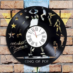 Può includere: Orologio in vinile nero con una silhouette di Michael Jackson che esegue la sua mossa di danza caratteristica. Il quadrante dell'orologio è bianco con indici orari in numeri romani neri e una lancetta delle ore rossa. L'orologio è circondato da un disco in vinile nero con il testo "KING OF POP" e "Michael Jackson" in lettere bianche.