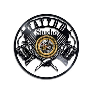 May include: Black vinyl record clock with a tattoo studio design. The clock features two crossed tattoo machines with the words "Tattoo Studio" and Roman numerals around the clock face.