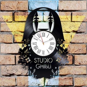 May include: A black vinyl record clock featuring a silhouette of No Face from the Studio Ghibli film Spirited Away. The clock face is white with black Roman numeral markings and a red hour hand and black minute hand. The clock is surrounded by a black silhouette of flowers and vines.