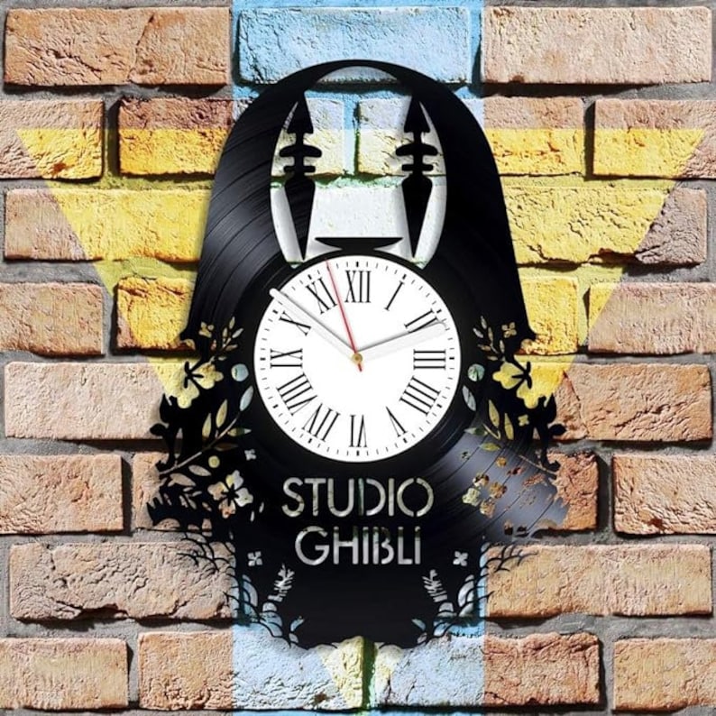 May include: A black vinyl record clock featuring a silhouette of No Face from the Studio Ghibli film Spirited Away. The clock face is white with black Roman numeral markings and a red hour hand and black minute hand. The clock is surrounded by a black silhouette of flowers and vines.