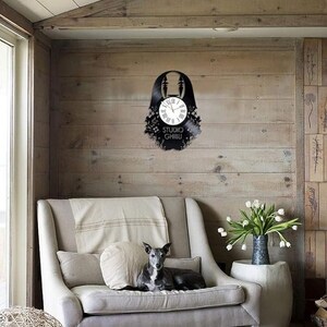 May include: A black vinyl record clock featuring a silhouette of a character from the Studio Ghibli film Spirited Away. The clock is hanging on a wood wall in a living room setting.