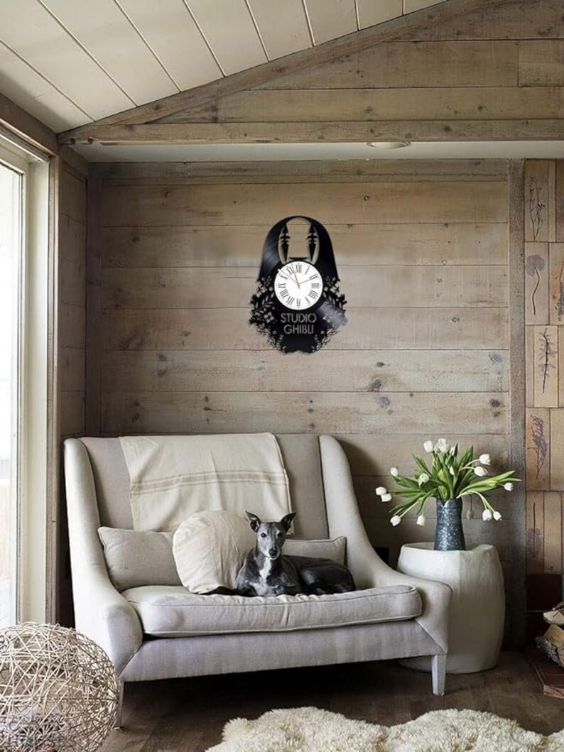 May include: A black vinyl record clock featuring a silhouette of a character from the Studio Ghibli film Spirited Away. The clock is hanging on a wood wall in a living room setting.