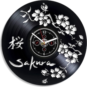 May include: Black vinyl record clock featuring a white floral design and the Japanese word "Sakura" in white. The clock face is black with white numbers and hands.