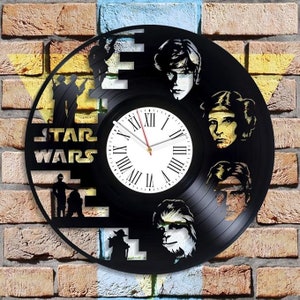 May include: A black vinyl record clock featuring characters from Star Wars. The clock face is white with Roman numerals and black hands. The characters include Luke Skywalker, Princess Leia, Chewbacca, and Yoda.