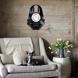 May include: A black vinyl record clock featuring a silhouette of a character from the Studio Ghibli film Spirited Away. The clock is mounted on a wall with a wooden background. The clock face is white with black numbers and hands. The clock is surrounded by a black vinyl record with a black and white image of the character.