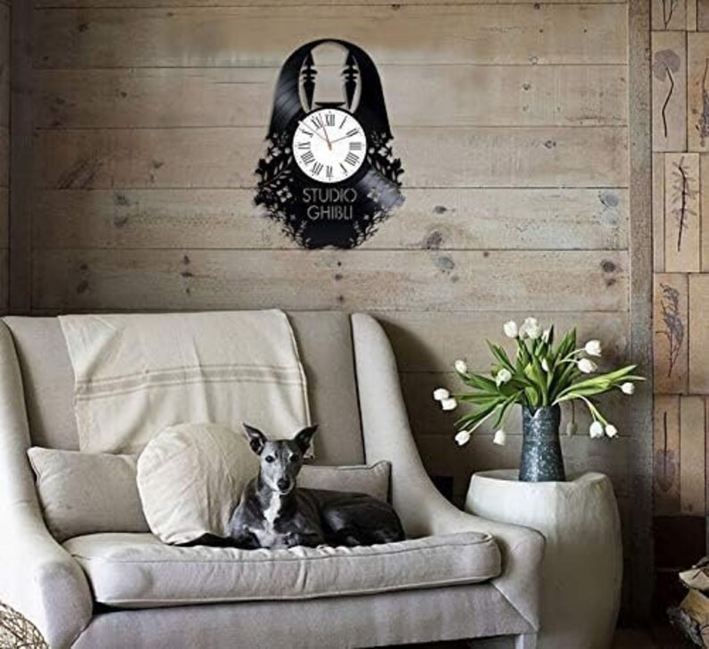 May include: A black vinyl record clock featuring a silhouette of a character from the Studio Ghibli film Spirited Away. The clock is mounted on a wall with a wooden background. The clock face is white with black numbers and hands. The clock is surrounded by a black vinyl record with a black and white image of the character.