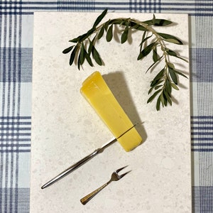 May include: A white rectangular serving board with a speckled surface. A wedge of yellow cheese is on the board with a silver cheese knife and a silver fork. A sprig of green olive branches is on the board.