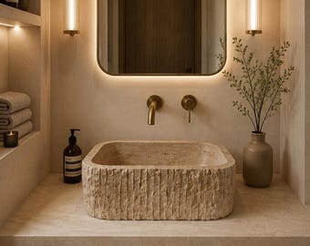 May include: A rectangular stone vessel sink with a textured, chiseled exterior finish sits on a matching stone countertop. The bathroom setup includes a wall-mounted brass faucet, a backlit rectangular mirror, and two vertical brass wall sconces.