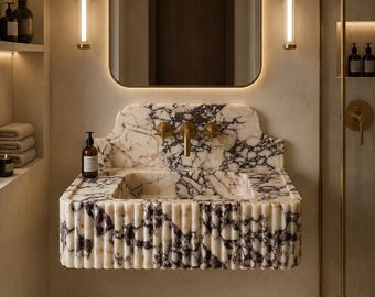 May include: A wall-mounted bathroom sink crafted from white and purple veined marble featuring a fluted front design. The sink includes a brass faucet and handles, positioned beneath a rectangular mirror with rounded corners and integrated lighting.