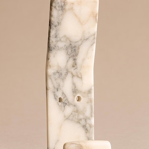 May include: A marble wall hook with a rectangular body and a curved hook at the bottom. The hook is white with grey veining and two small holes. The hook is designed for hanging items.