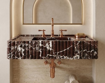 May include: A modern bathroom sink made of red marble with white veining. The sink has a copper faucet and fixtures. A mirror with a gold frame is above the sink. Rolled towels are in a wicker basket below the sink.