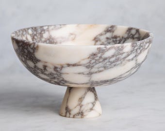 May include: A white marble pedestal bowl with gray veining. The bowl has a rounded shape and sits on a short, conical base. The marble has a natural, patterned appearance, suitable for serving or display.